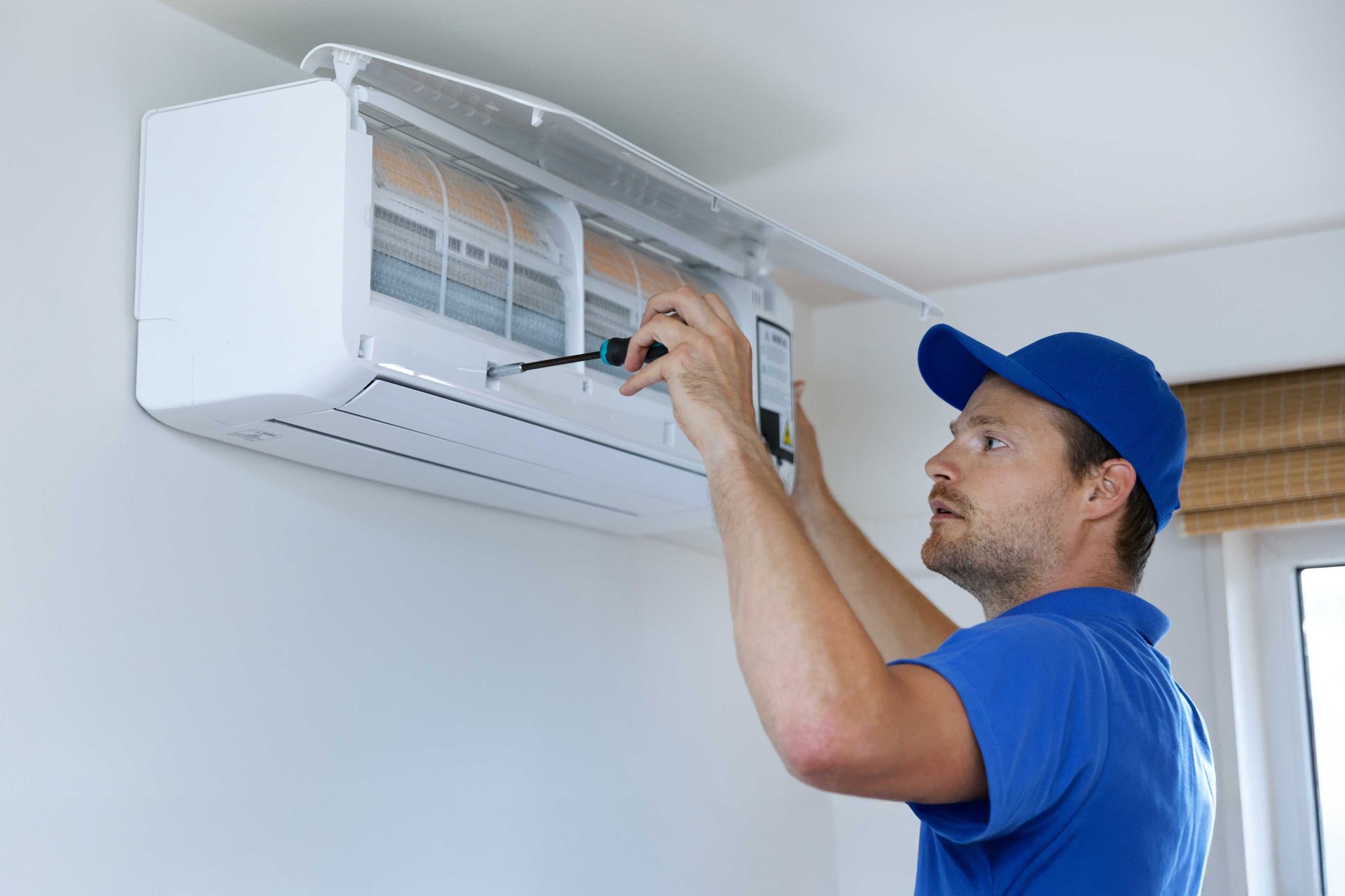 HVAC technician in blue uniform, fixing a wall-mounted air conditioner with a screwdriver. White room setting.