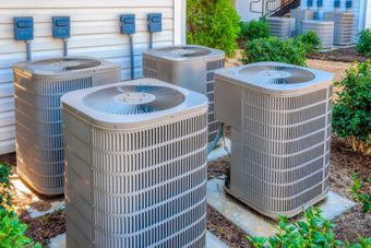 Four gray air conditioning units outside a building with electrical boxes and landscaping. Four gray air conditioning units outside a building with electrical boxes and landscaping.
