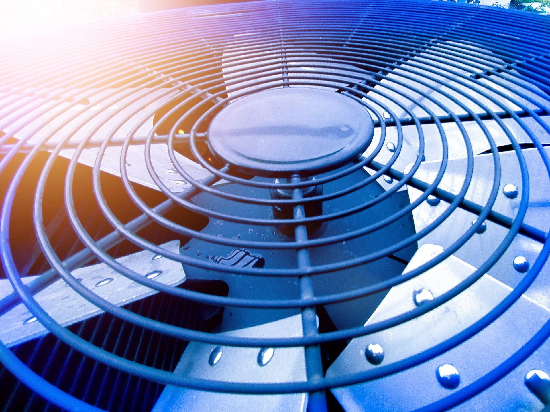 Close-up of an air conditioner fan covered by a protective metal grill; blue tones.