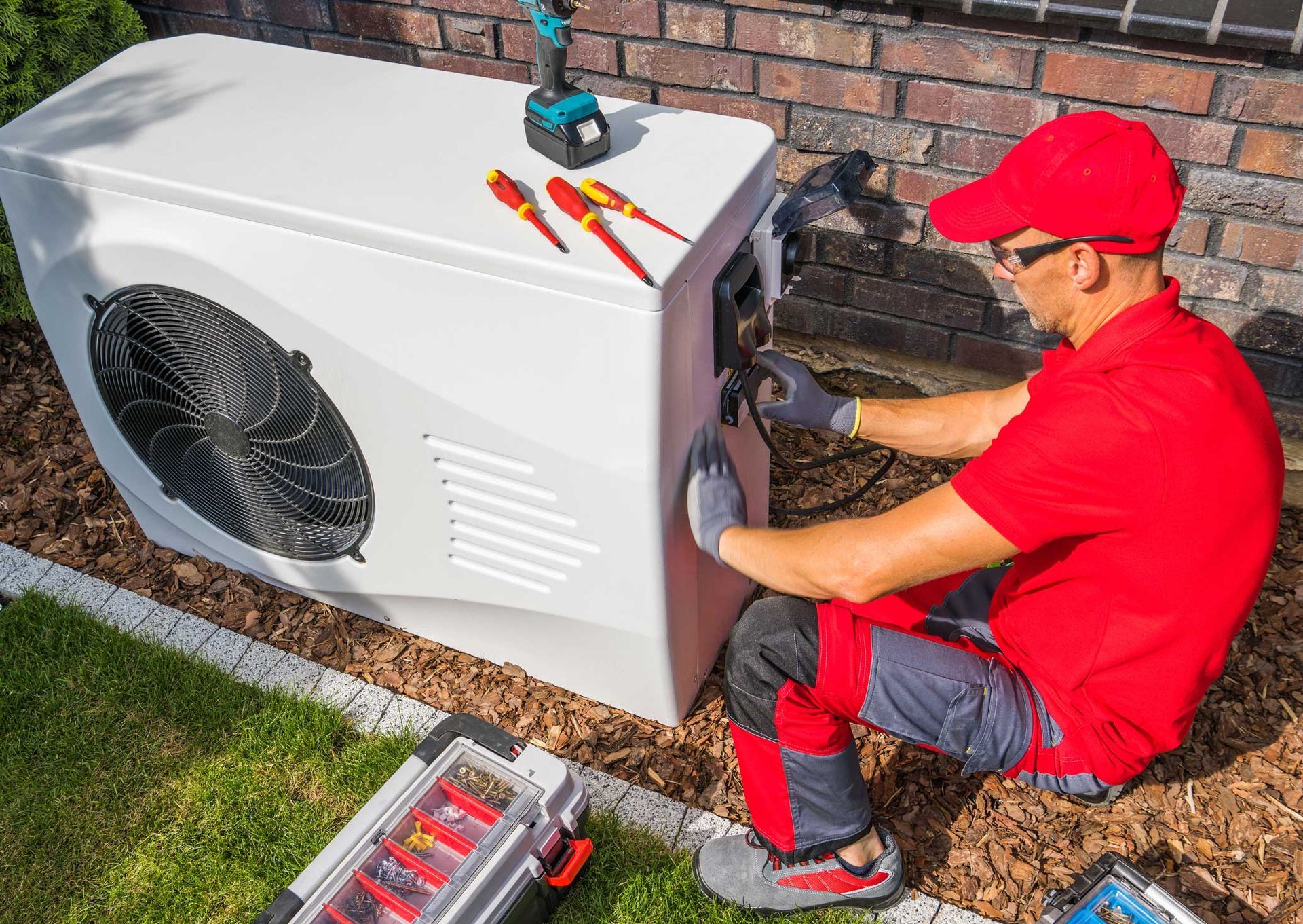 A technician in red works on a white HVAC unit outdoors, using tools.