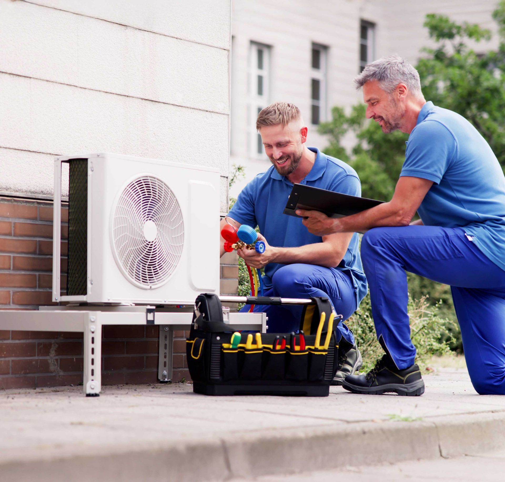 Two technicians inspecting an outdoor HVAC unit, one kneeling and working, the other holding a clipboard.