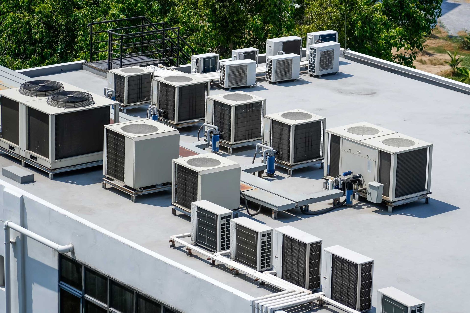 Air conditioning units on a building rooftop. Gray and white units with trees in the background.