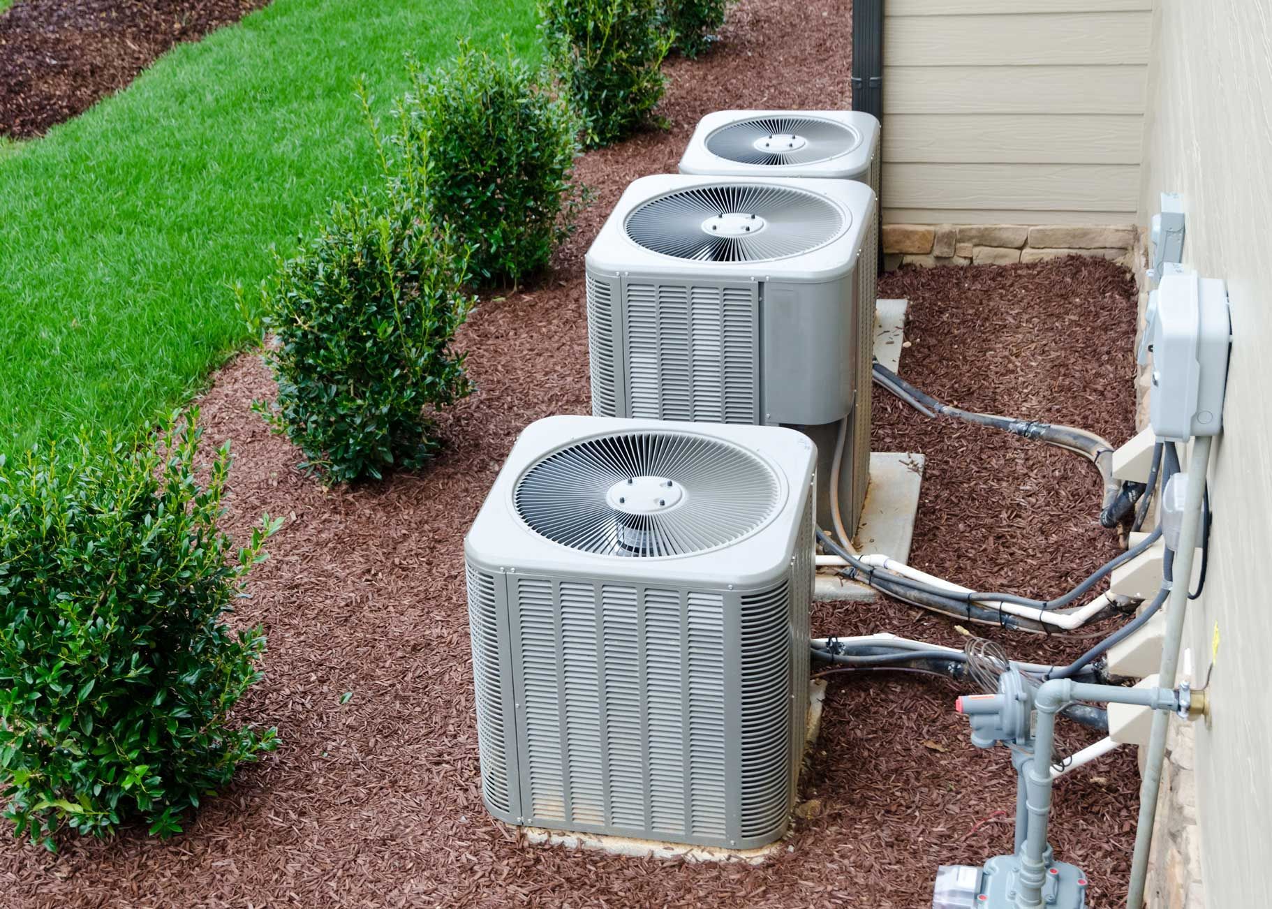 Three air conditioning units on gravel next to a building, with greenery in the background.