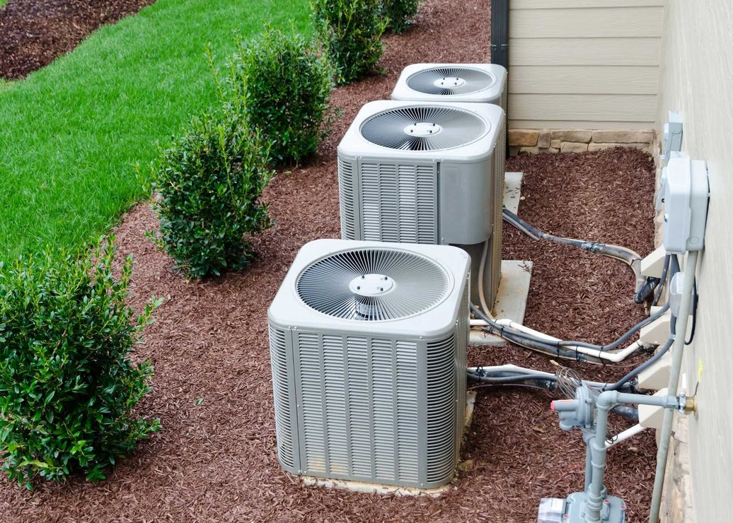 Three air conditioning units on gravel next to a building, with greenery in the background. Three air conditioning units on gravel next to a building, with greenery in the background.