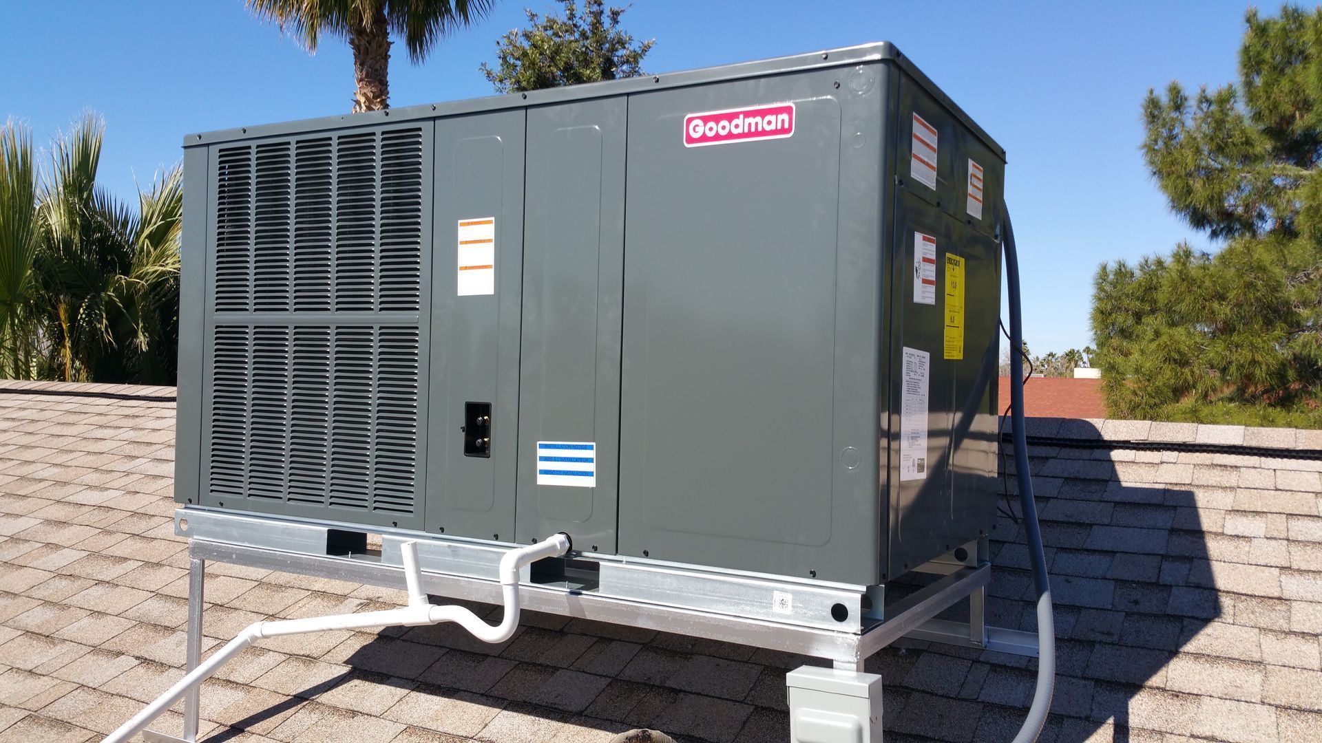 A gray Goodman HVAC unit on a rooftop platform, against a blue sky with trees.