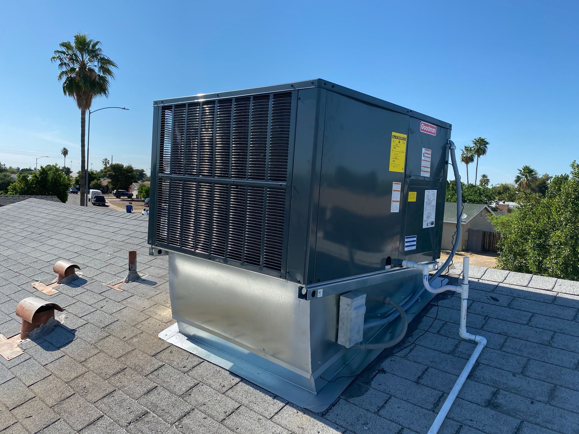 Rooftop HVAC unit on a shingled roof under a clear, blue sky with palm trees in the background.
