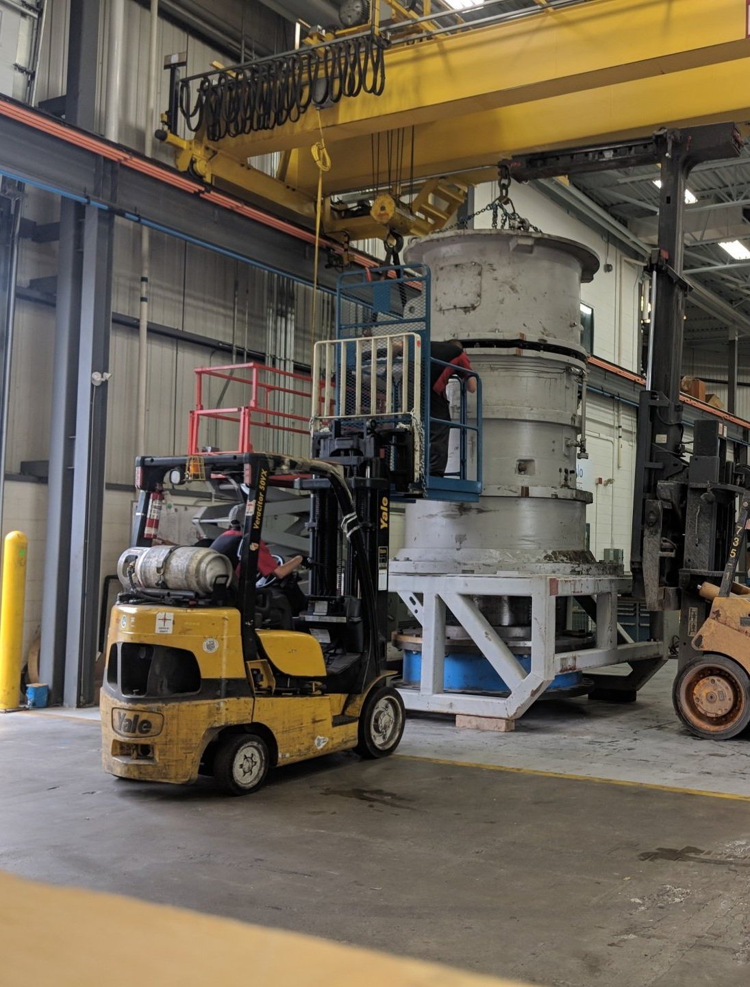 A yellow forklift in a warehouse with a yellow crane in the background