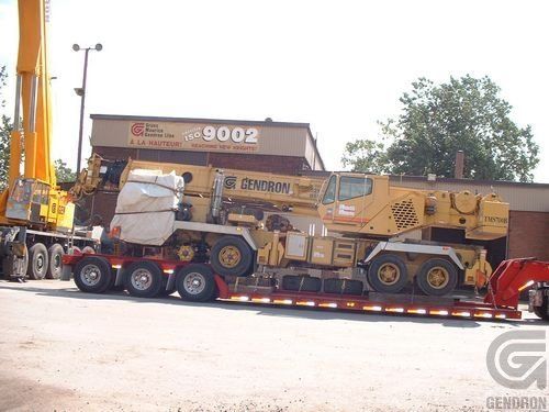 Une grande grue jaune est sur une remorque devant un bâtiment qui indique 9002