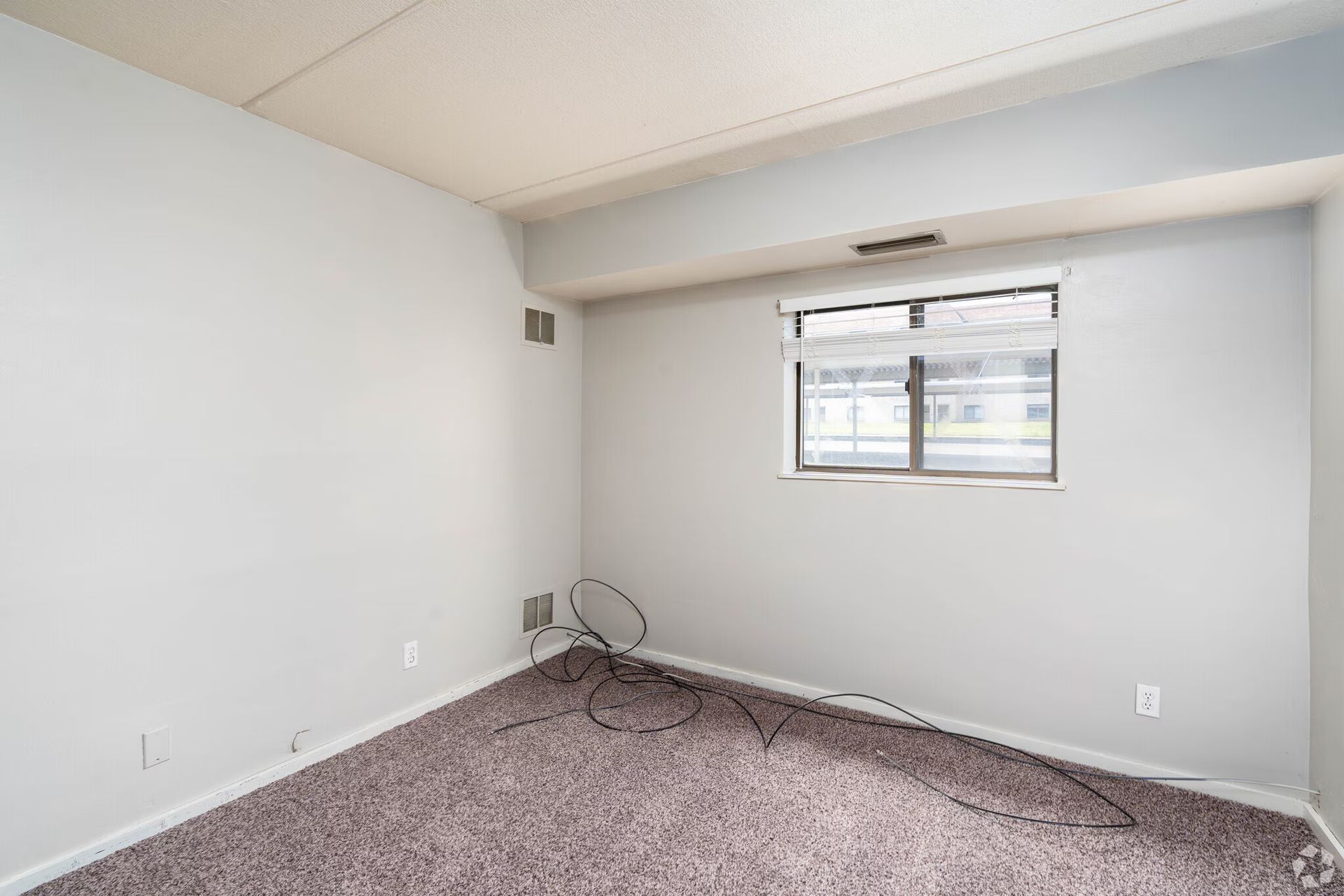Empty bedroom with gray walls, a window, and brown carpet. Wires are visible on the floor.