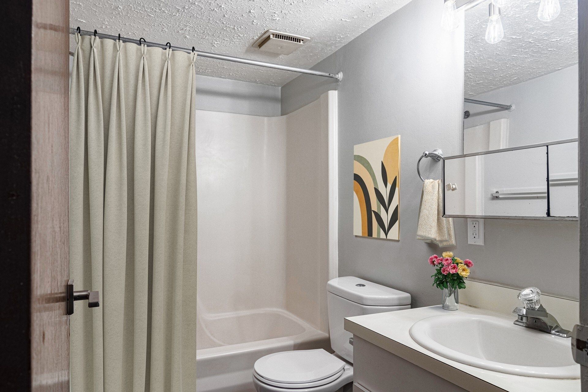 Bathroom with shower/tub, toilet, sink, mirror, and decorative art; light gray walls and beige shower curtain.