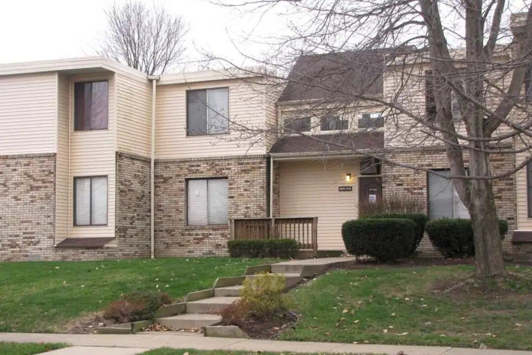 Two-story townhouses with beige siding, stone accents, and front entry stairs. Green lawn and leafless trees.