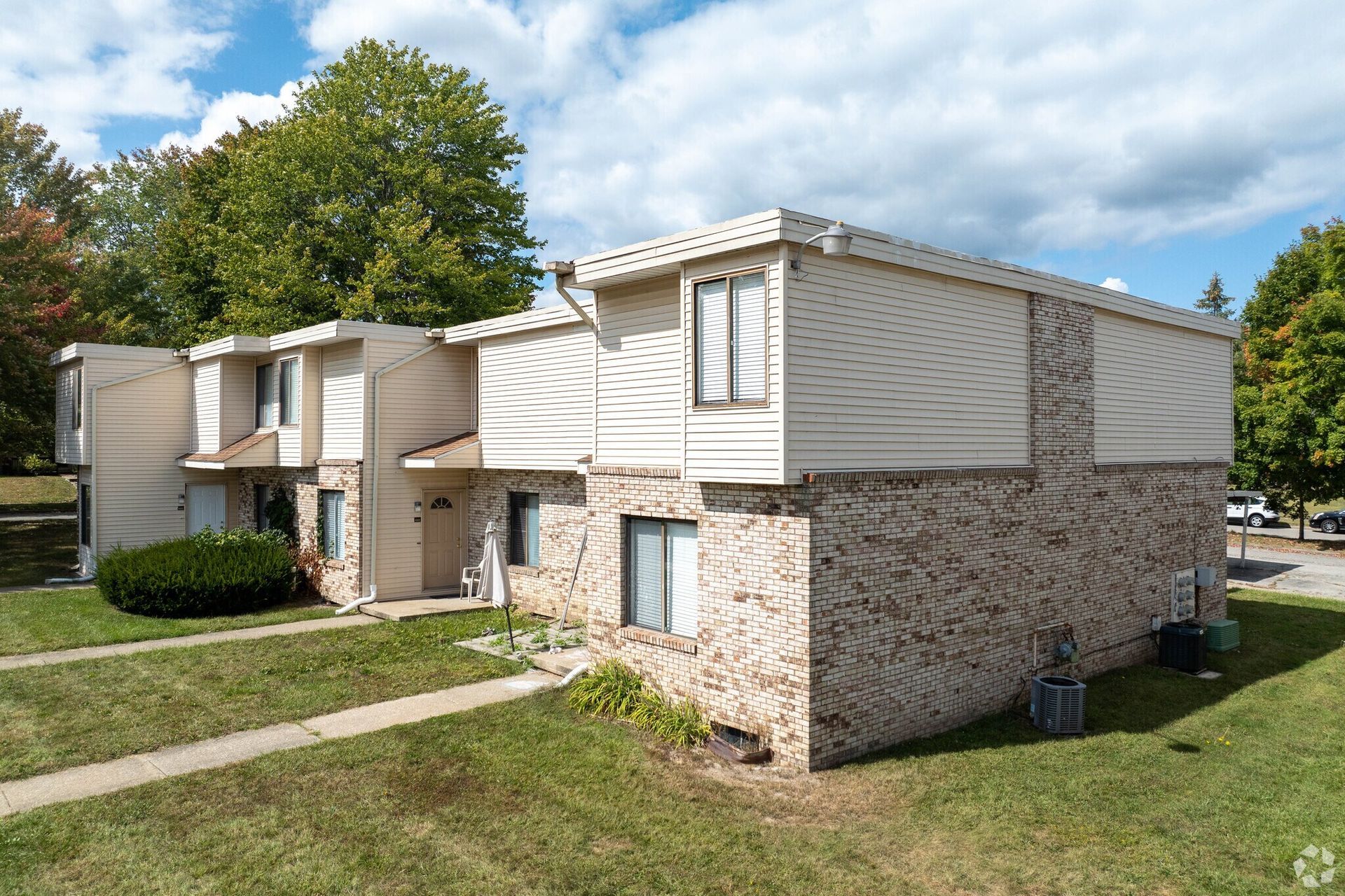 Two-story tan and brick apartment building with green lawn, trees, and blue sky.