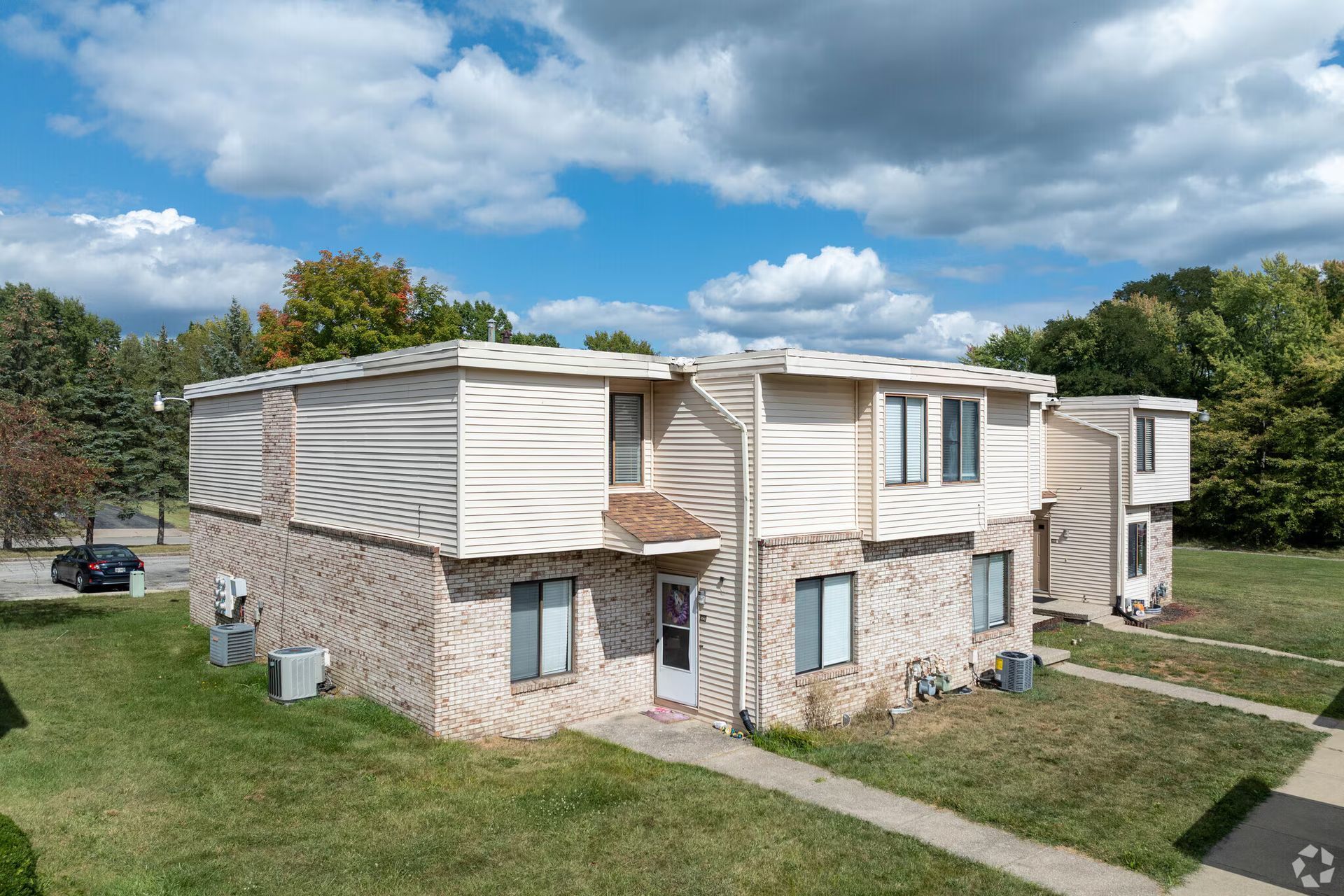 Two-story brick and siding apartment building with green lawn, trees, and cloudy sky.