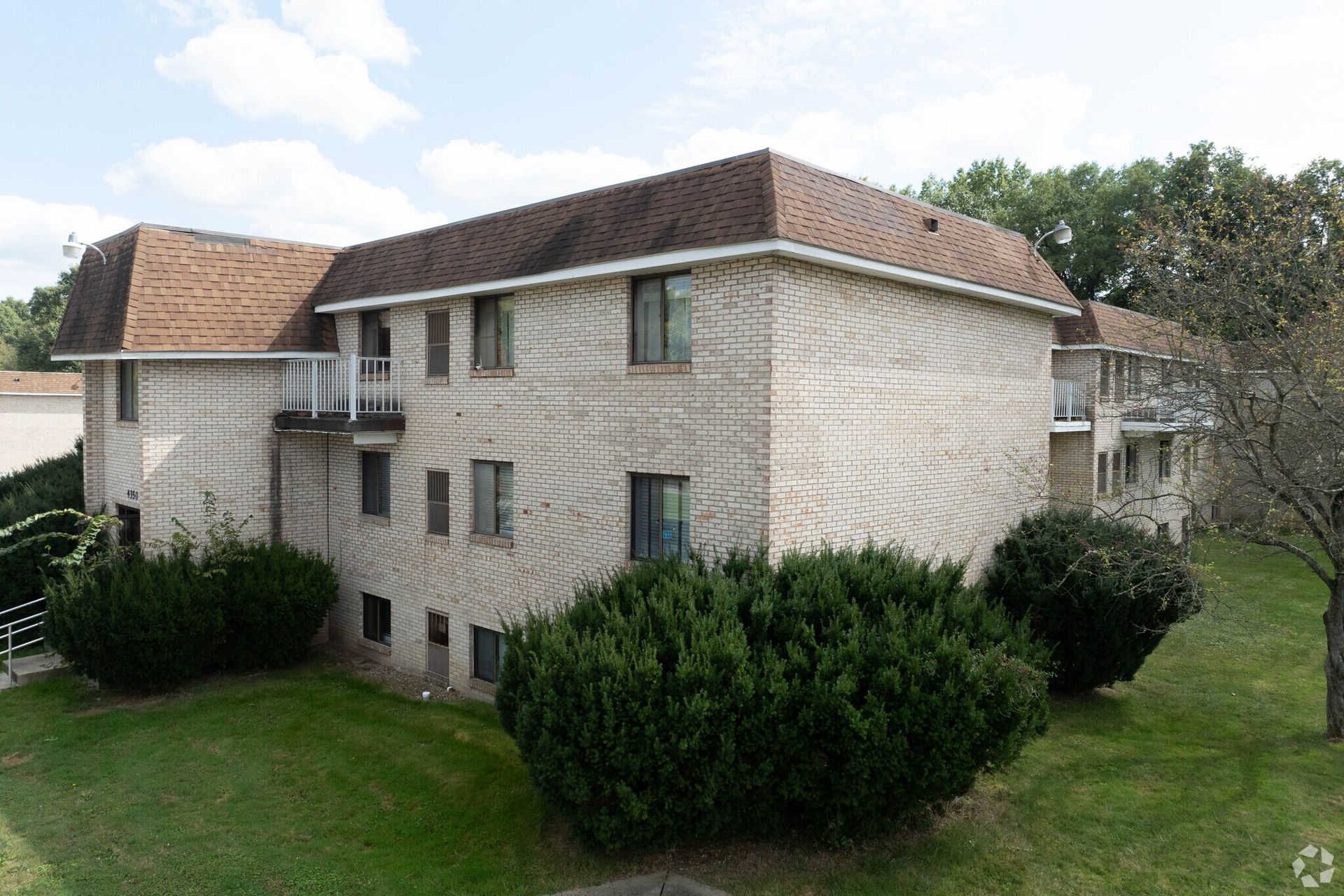 Two-story brick apartment building with brown roof and green bushes in front.