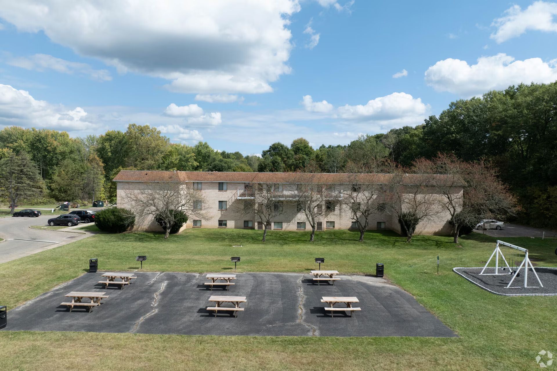 Two-story apartment building with picnic tables in front, playground to the right, and trees in background.