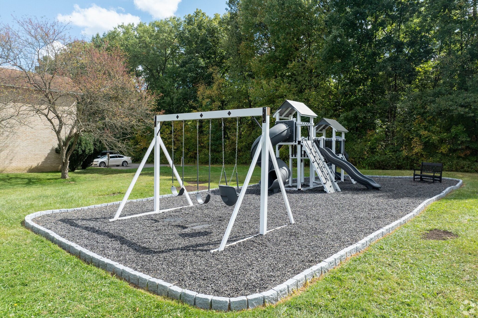 Playground with swings and slide on a gravel bed, bordered by grass and trees.