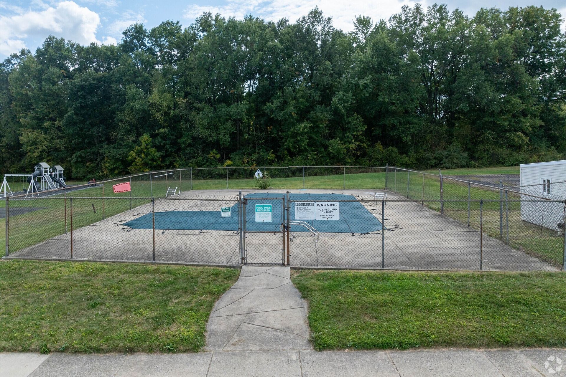 A gated, fenced-in outdoor swimming pool covered with blue tarp, surrounded by grass and trees.
