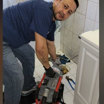 A man in blue shirt and jeans uses a plumbing snake on the bathroom floor. He's wearing gloves, working near a white cabinet.