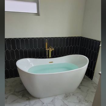 A white, oval-shaped bathtub filled with water sits in a modern bathroom. Black tile wall with gold faucet.