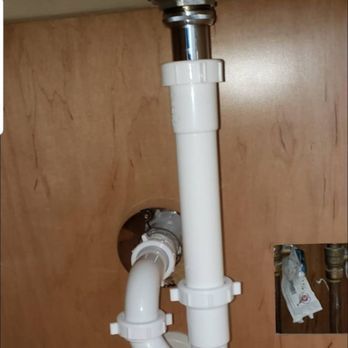 White plumbing pipes under a cabinet, connected to a silver drain. The pipes are angled and curved.