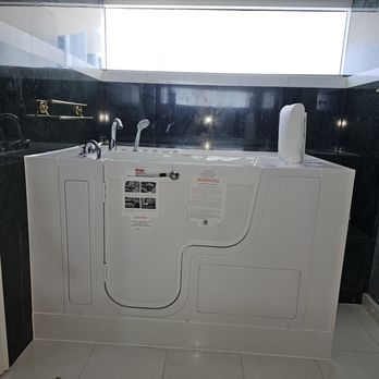 A white walk-in bathtub with a door and a raised seat. It's in a bathroom with dark tile and a window above.