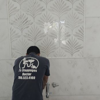 A man in a grey t-shirt with a logo works on a faucet below a decorative tile wall. The wall has white and grey leaf-patterned tiles.