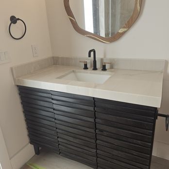 Dark wood bathroom vanity with a white countertop, black faucet, and a free-form mirror. A black towel ring is on the wall.