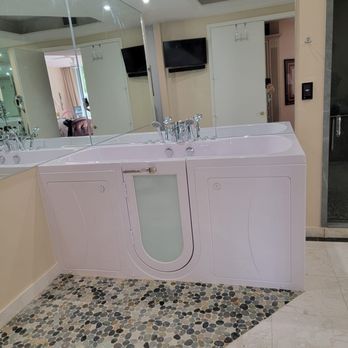 A walk-in bathtub with a glass door, chrome fixtures, and a pebble floor mat in a bathroom with a mirror.