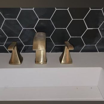 Brushed gold bathroom faucet with three handles against a black hexagonal tiled backsplash and a white sink.