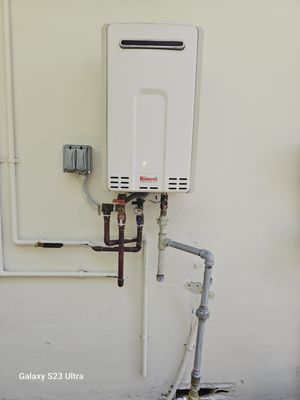 A white, wall-mounted Rinnai tankless water heater with copper and grey pipes on a light-colored wall. Electrical box present.