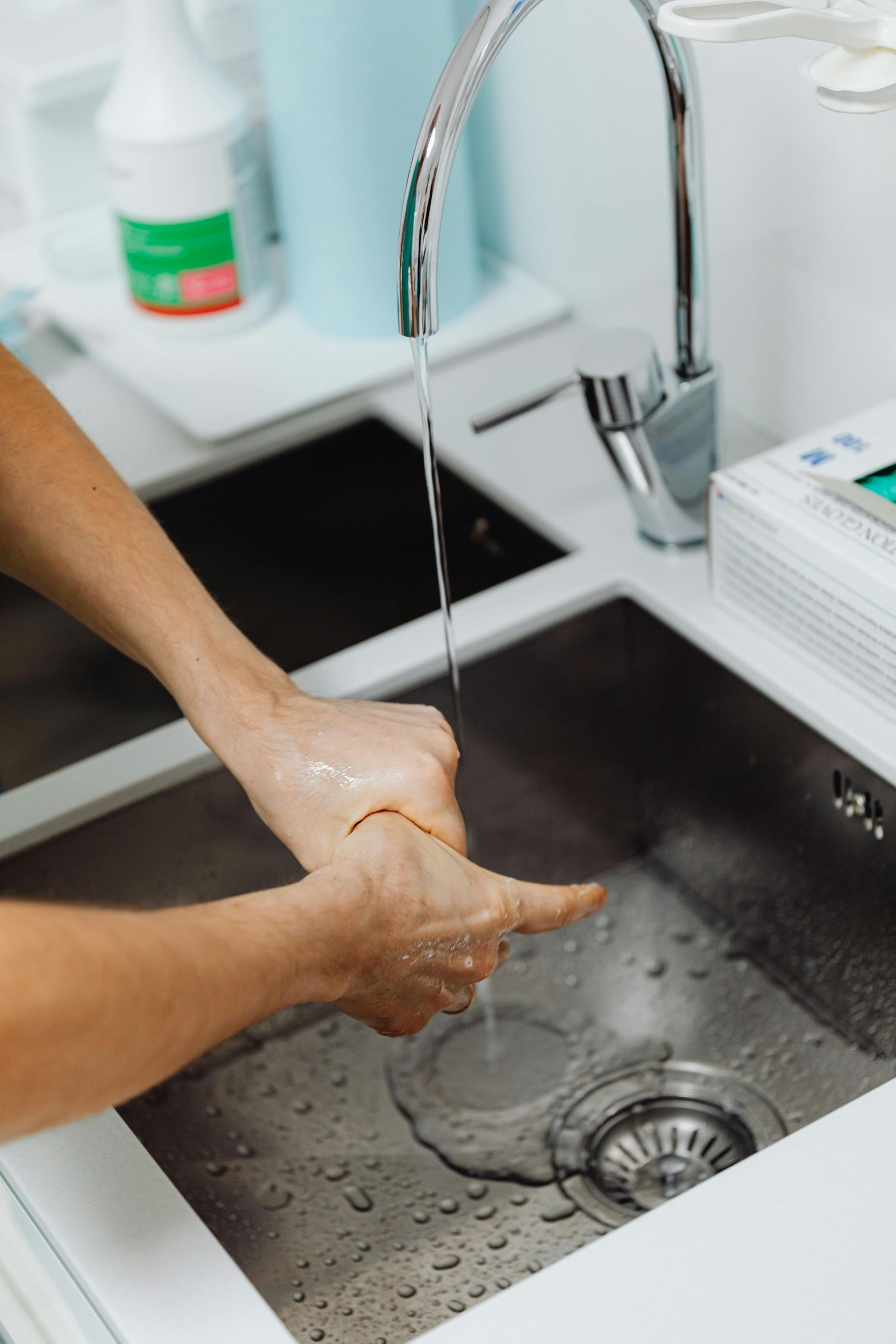 Person washing hands in a stainless steel sink under running water in a medical or clinical setting.