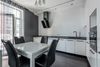 Modern white kitchen with a dining table, black chairs, and stainless steel appliances. Dark accent wall and sheer curtains frame the balcony.