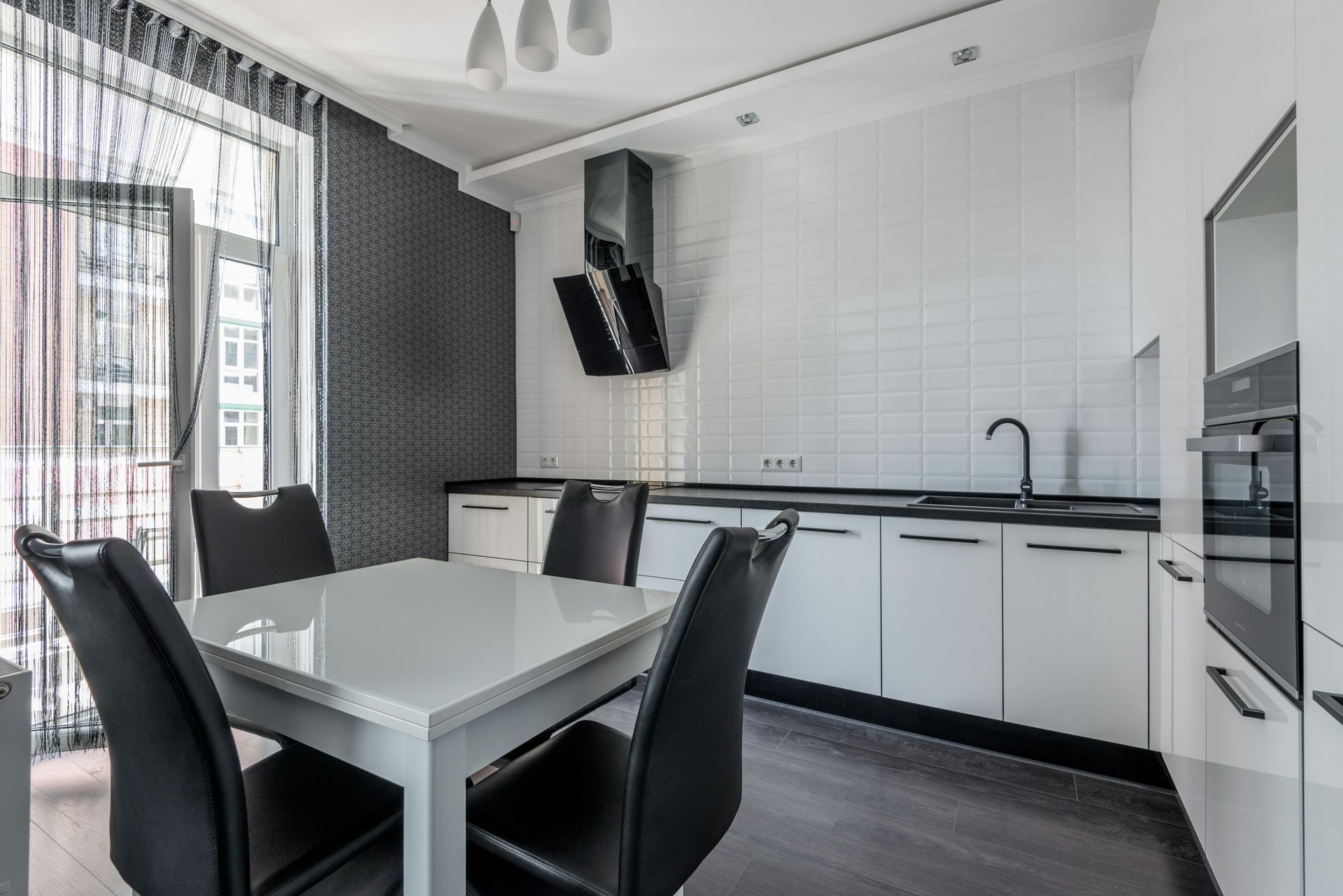 Modern white kitchen with a dining table, black chairs, and stainless steel appliances. Dark accent wall and sheer curtains frame the balcony.