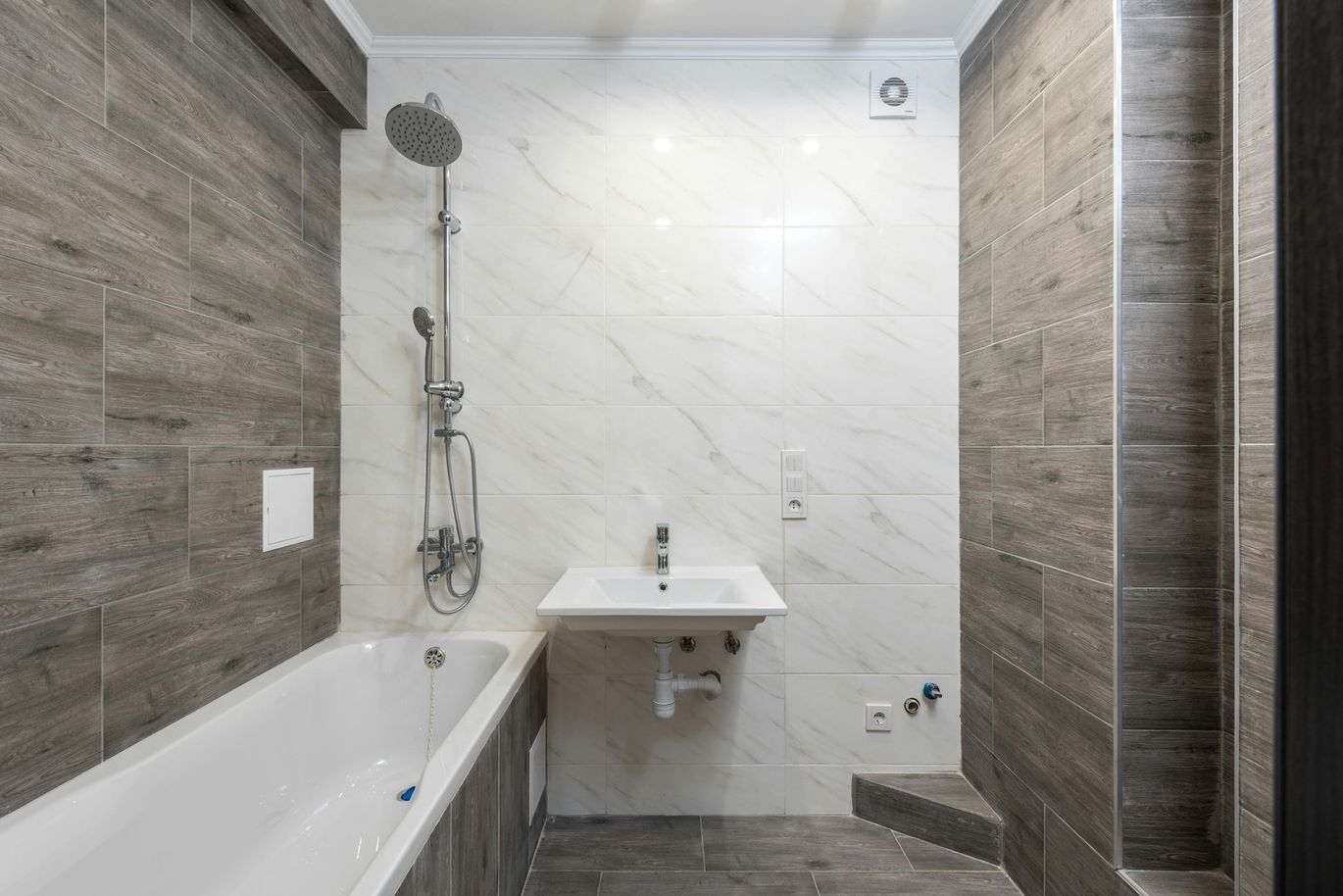 Modern bathroom with white bathtub, sink, and shower fixtures against gray and white tiled walls.