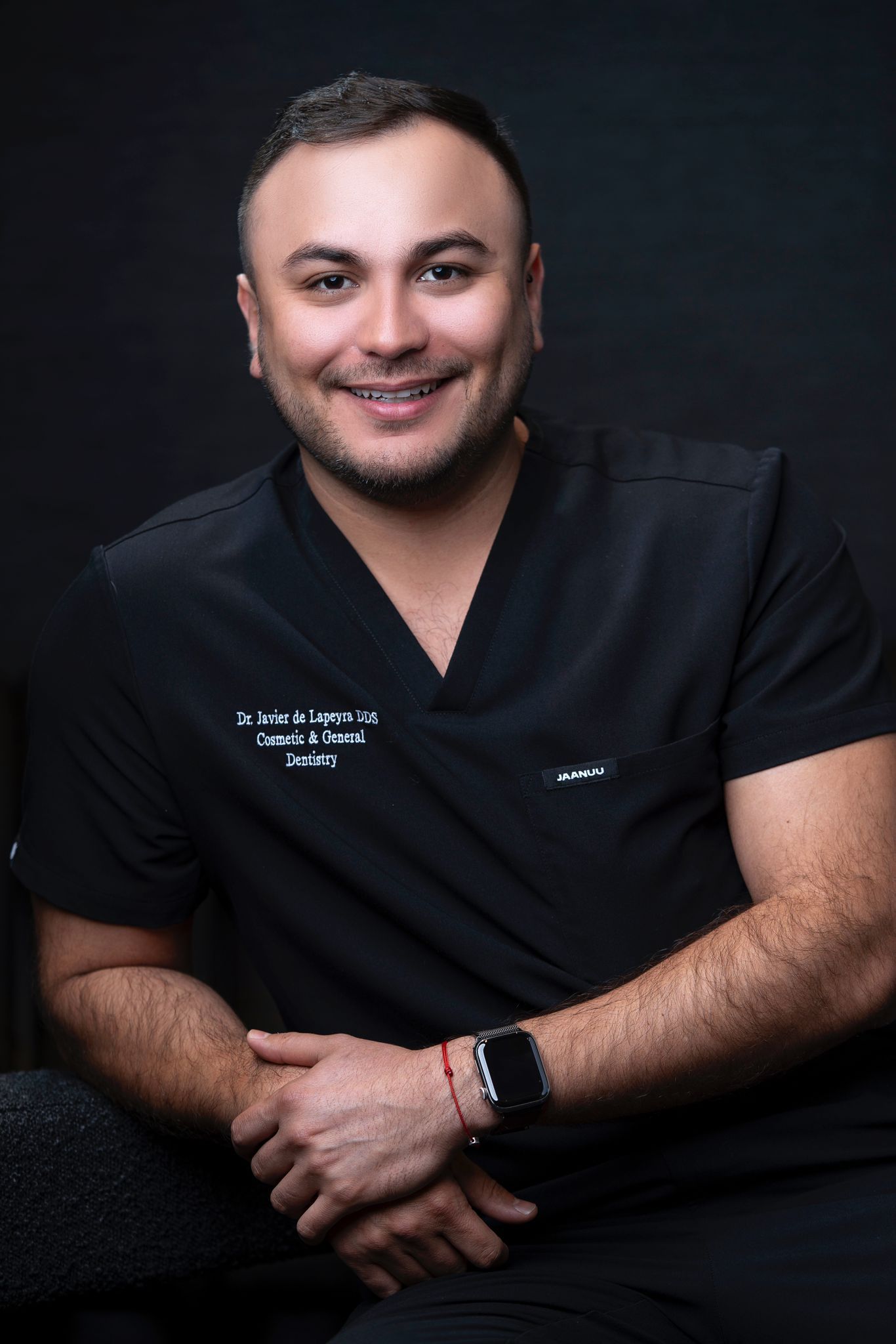 A man in a black scrub top is sitting with his hands folded and smiling.