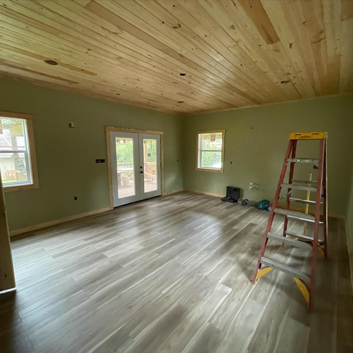 Room under construction: wood ceiling, green walls, wood-look floor, French doors, windows, ladder.
