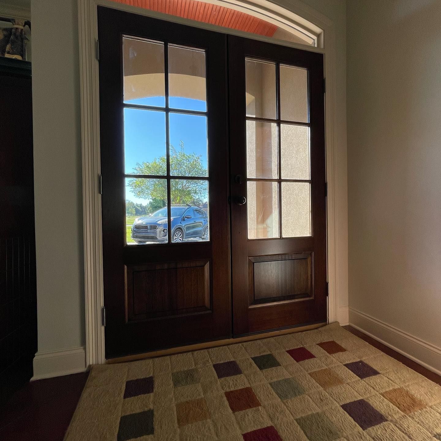Dark wood double doors with glass panels, leading to an outside view of a car and trees. Rug in front.