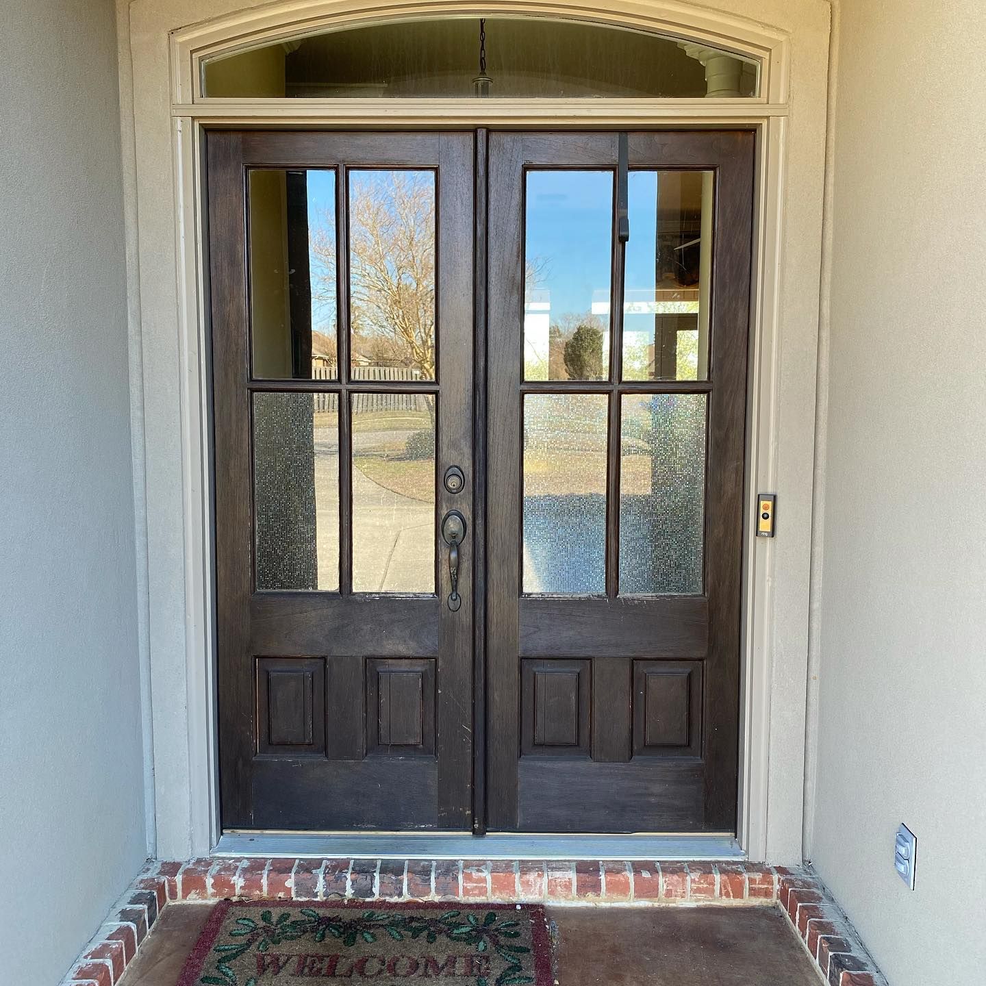 Double brown front doors with glass panels, set in a neutral-toned doorway with a welcome mat.