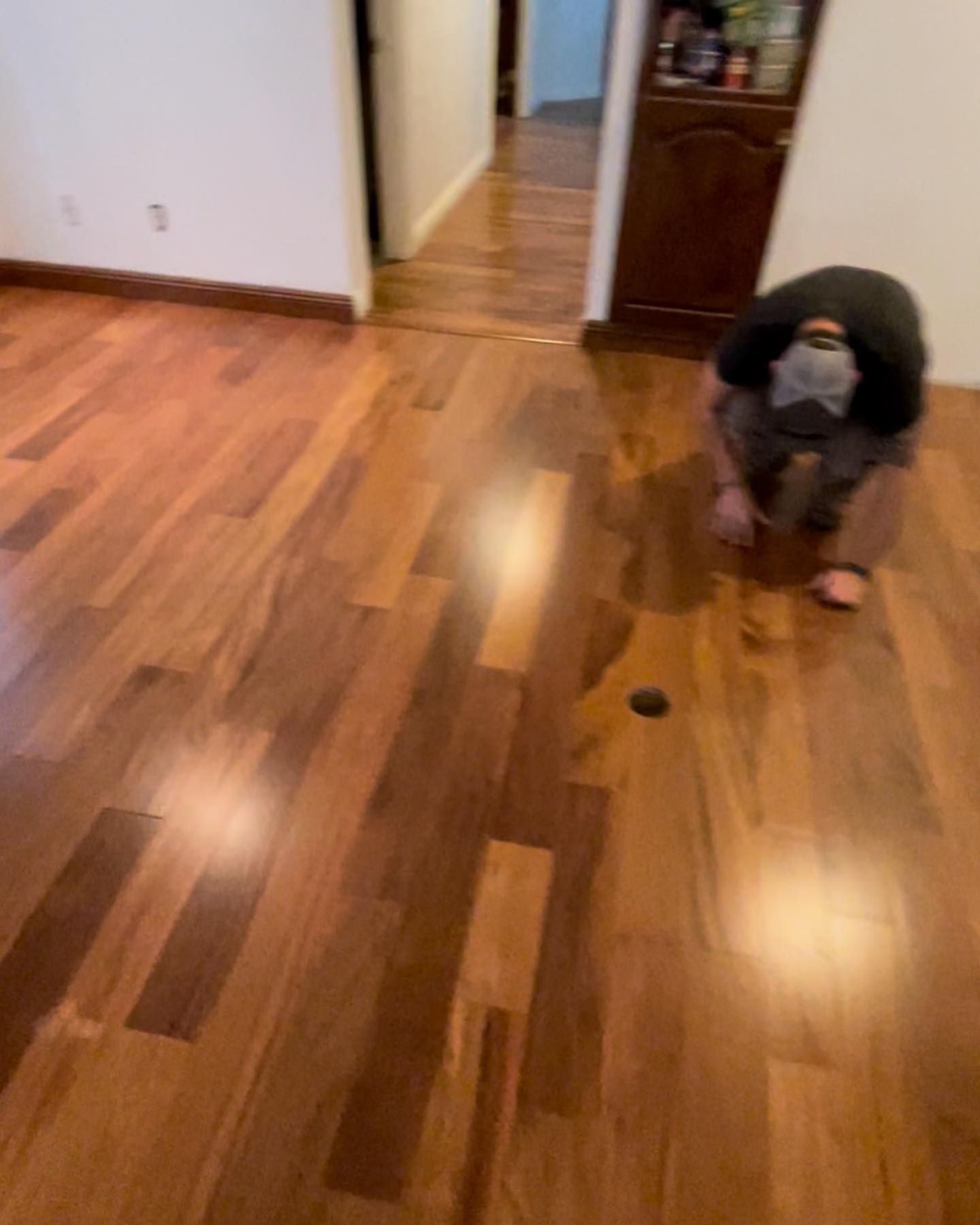 A person kneels on a hardwood floor. The floor is shiny and light brown. A dark hole is near the person.