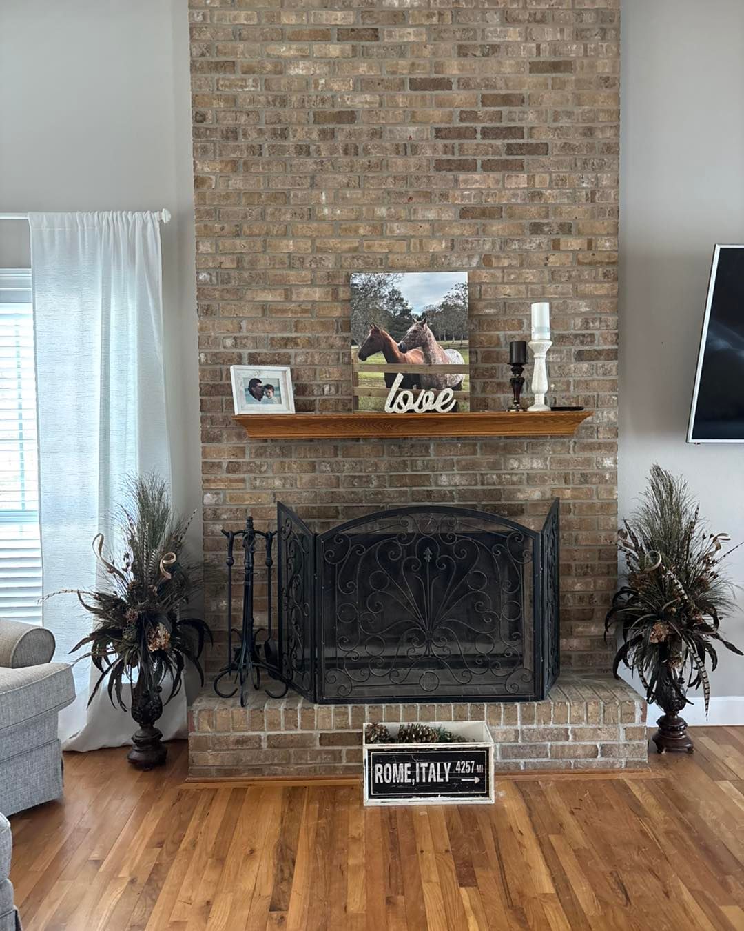 Fireplace with brick surround, wooden mantel holding art, flanked by tall floral arrangements, on hardwood floor.