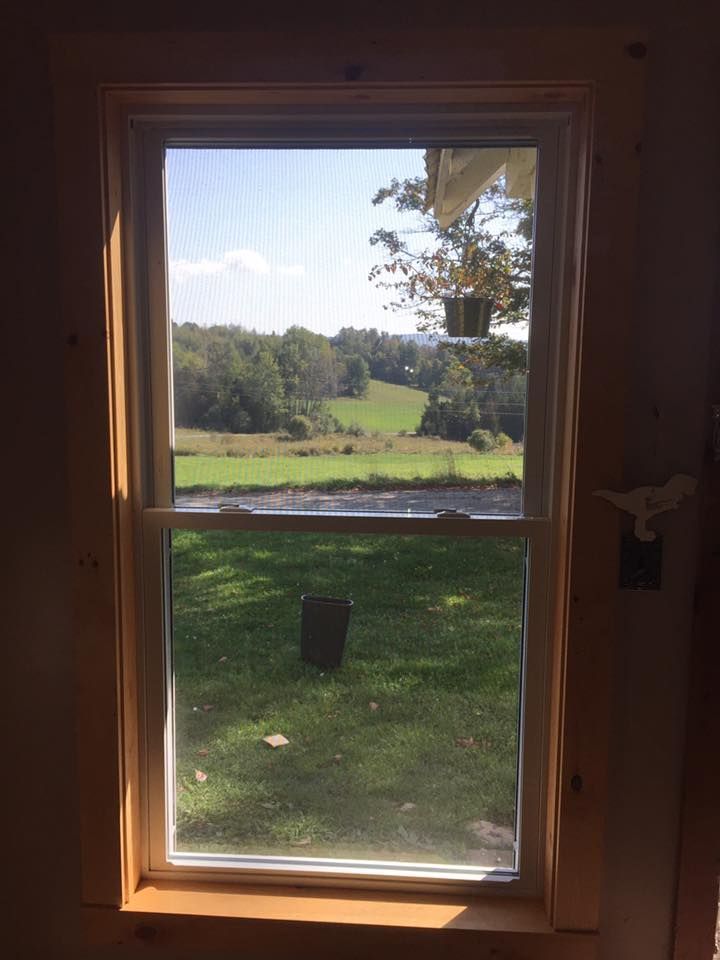 Window view of a green field, trees, and blue sky. A rectangular grave marker stands on the grass.