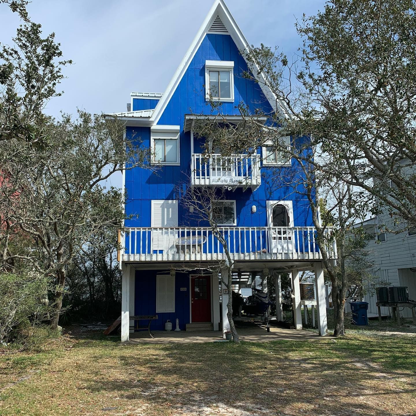 Bright blue beach house with white trim, built on stilts, surrounded by trees.