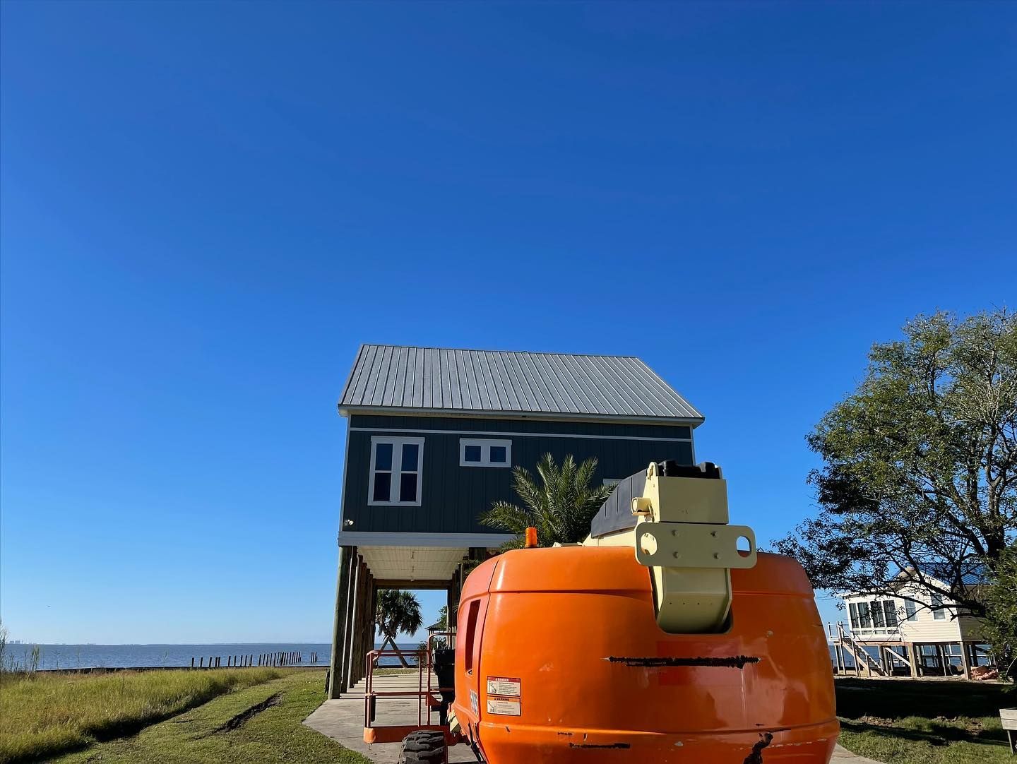 A teal house on stilts with an orange machine in front, against a clear blue sky and water.