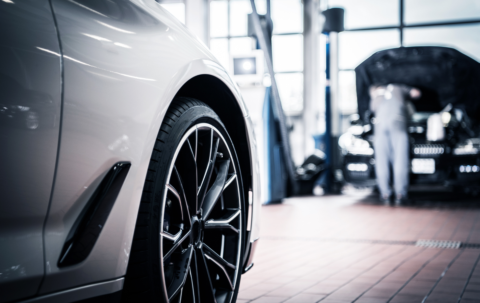 Silver car wheel in a garage with a mechanic working on a vehicle in the background.