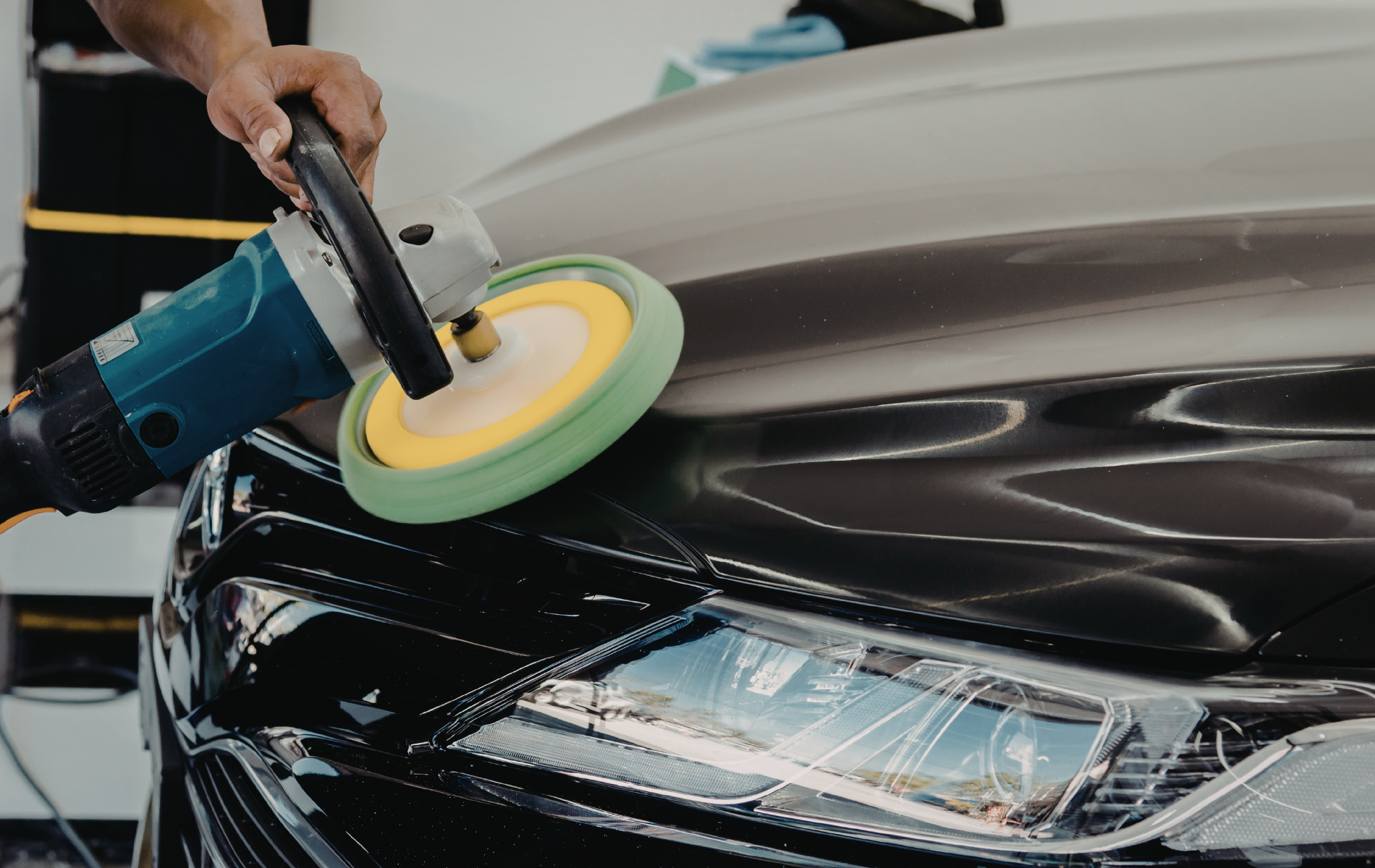 Person using a polisher on a black car hood, buffing a section of the vehicle.