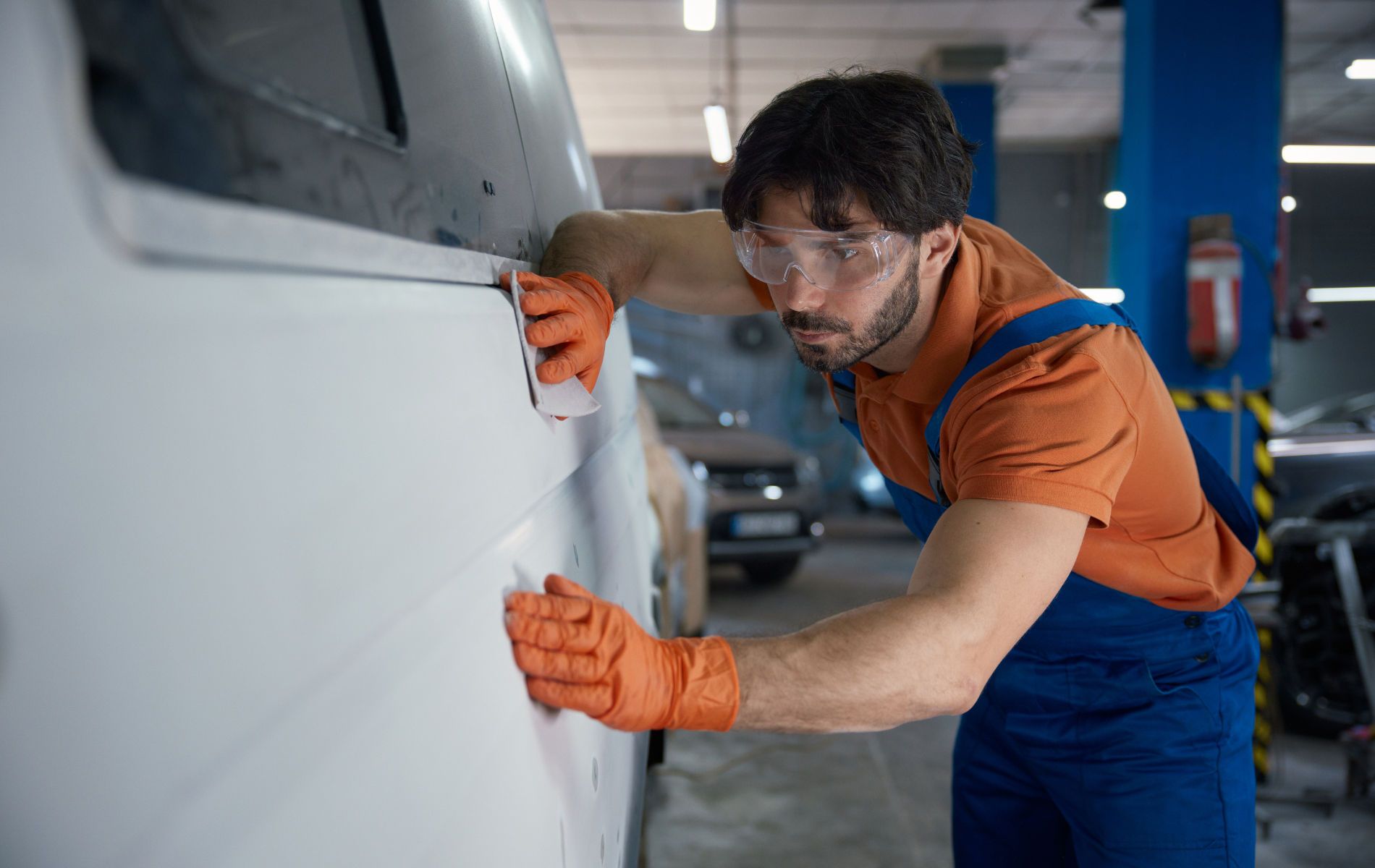 Mechanic sanding a white vehicle door, wearing safety glasses and orange gloves, in a garage.