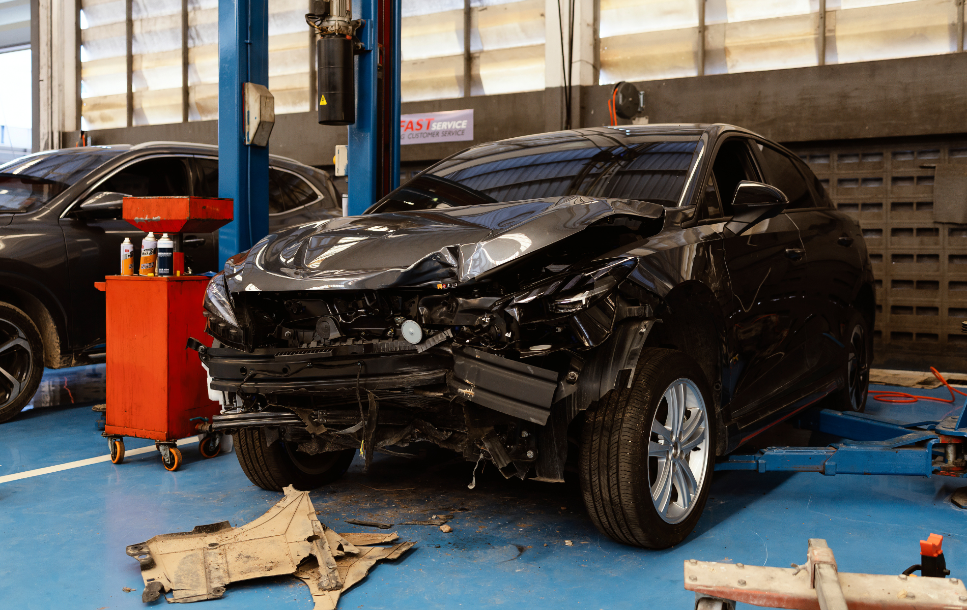 Damaged gray car in a repair shop; front end heavily wrecked; parts on the floor.