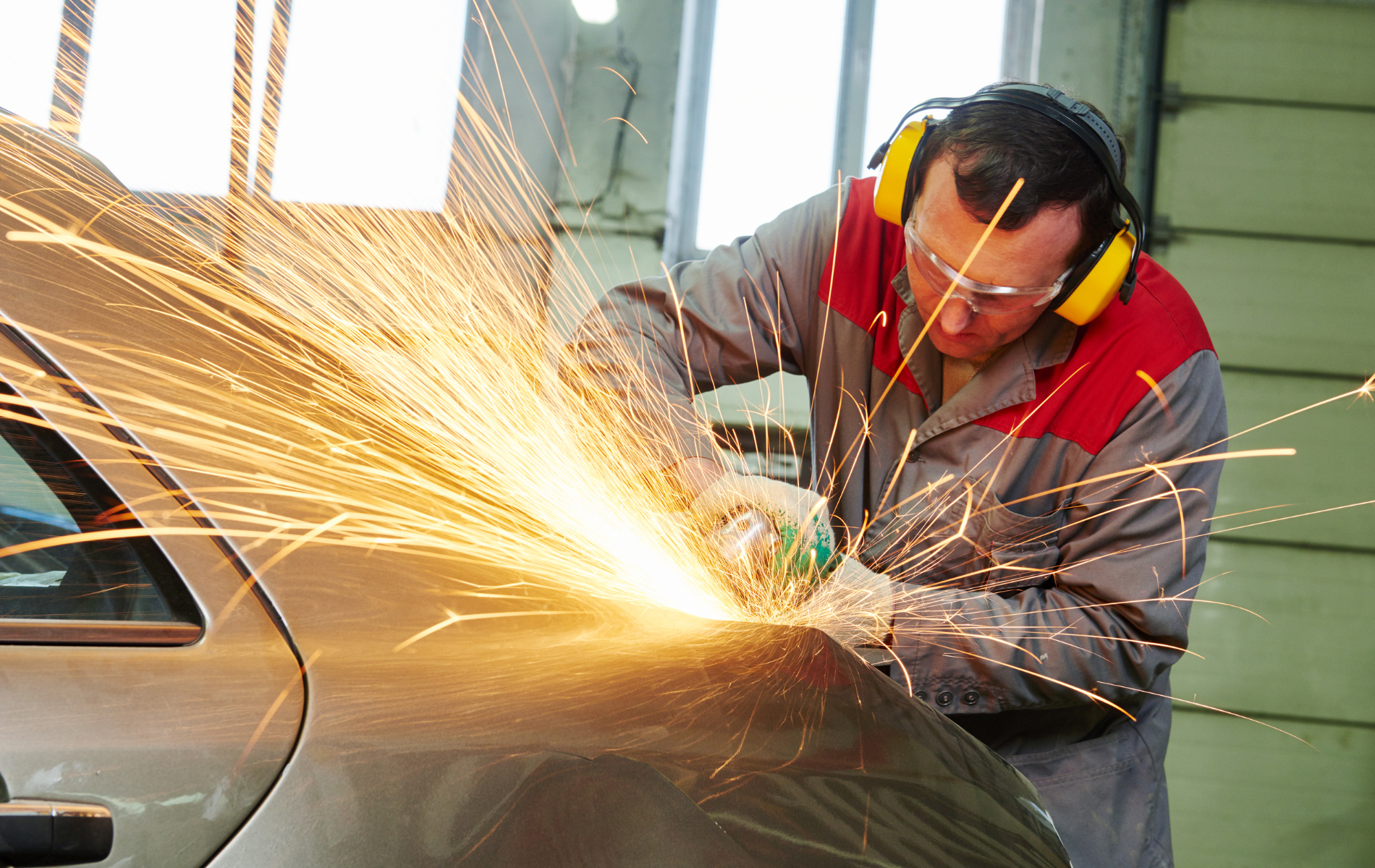 A mechanic grinds a car fender, creating sparks while wearing safety gear.