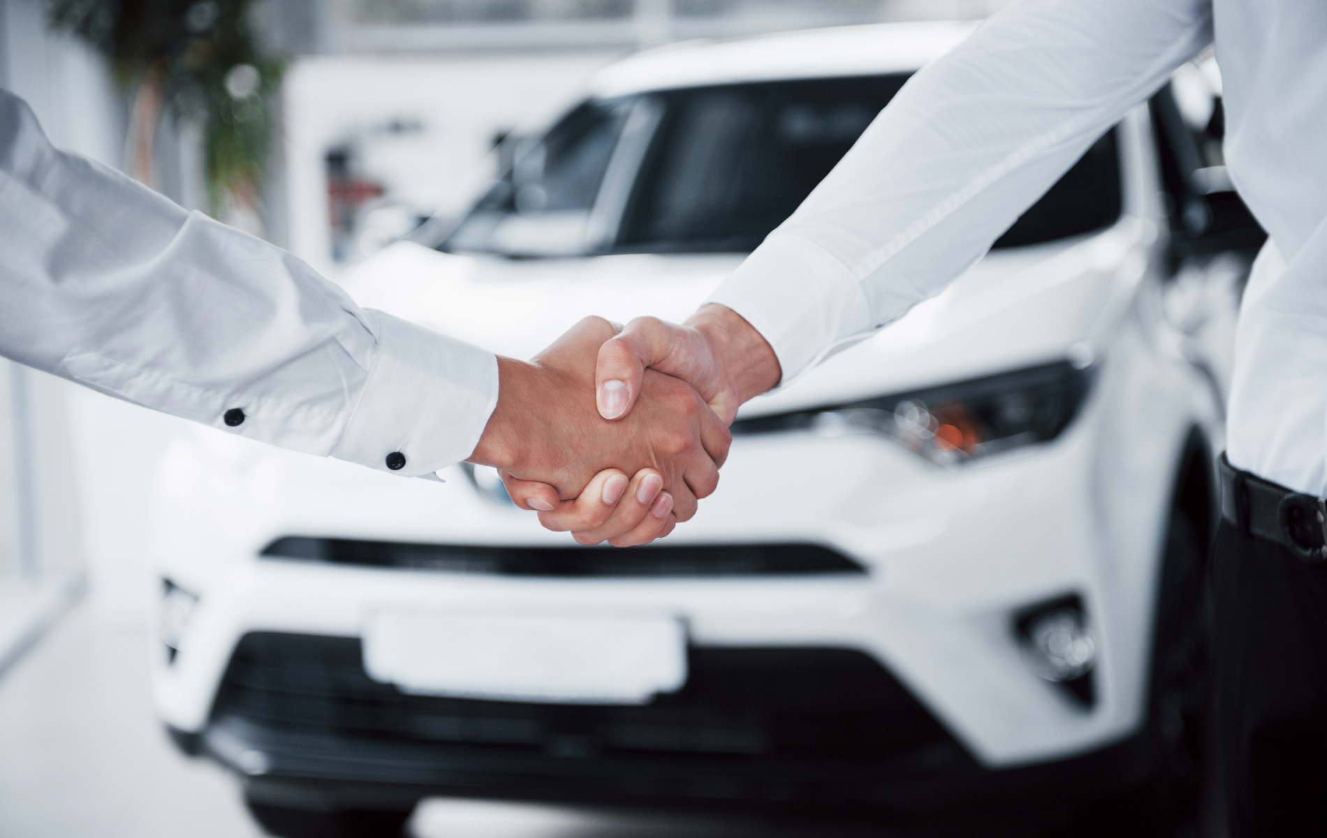 Two people shake hands in front of a white car in a showroom.