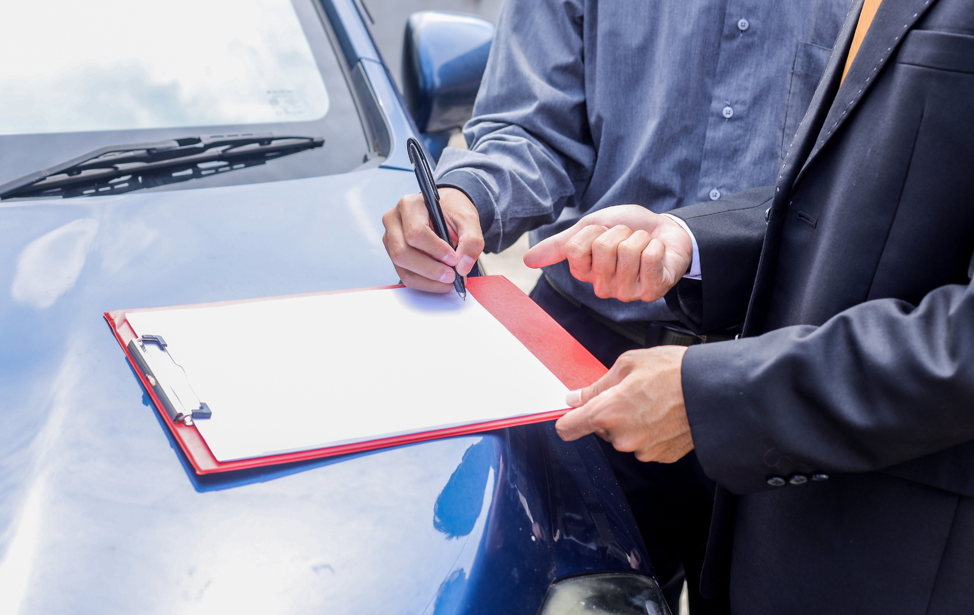 Person signing document on clipboard next to a blue car.